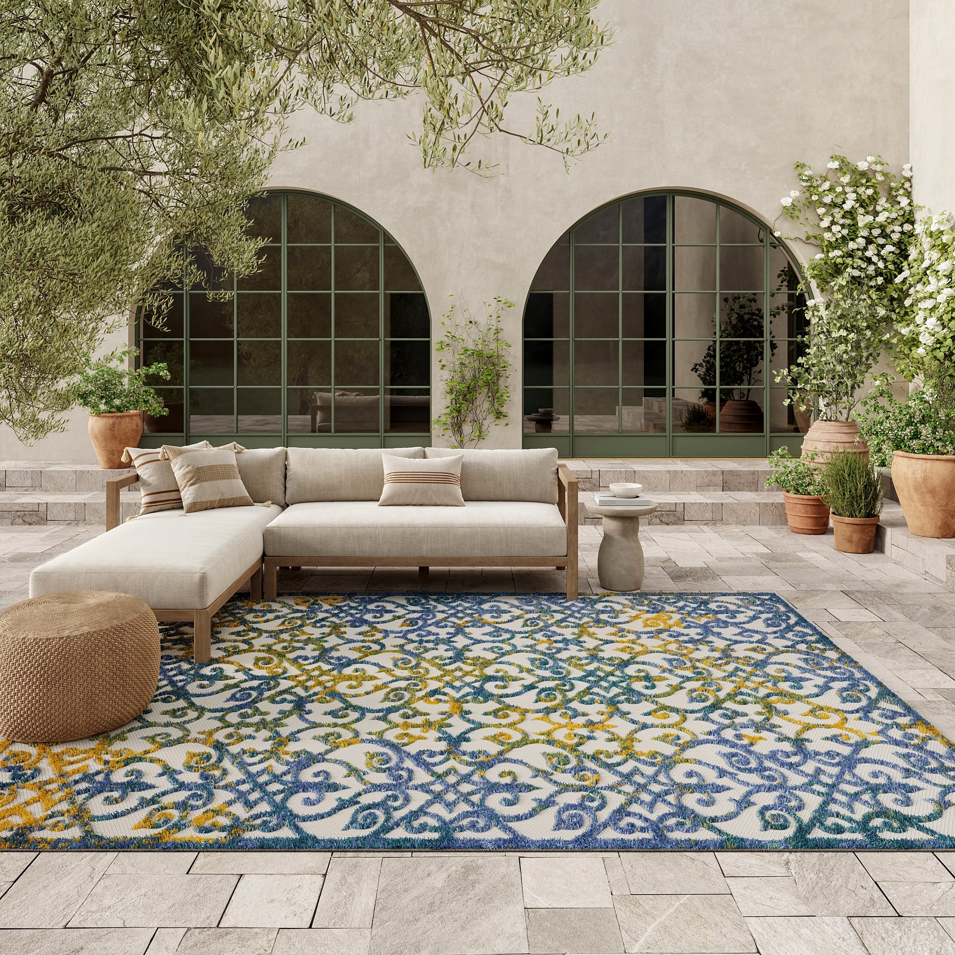 Outdoor courtyard scene with beige sectional sofa on a blue-yellow ornate rug, arched windows, potted plants.