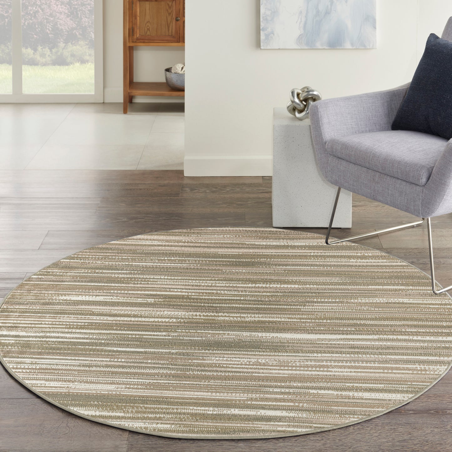 Round striped beige-taupe area rug on hardwood floor with gray upholstered lounge chair and navy cushion in a modern living room.