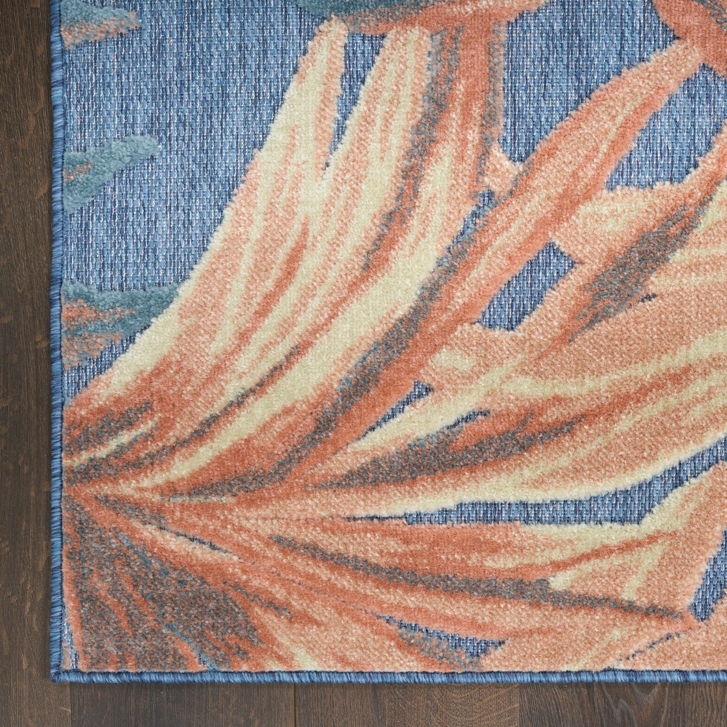 Decorative rug with a large orange coral leaf motif on a blue backdrop; blue edge stitching, placed on a wood floor.