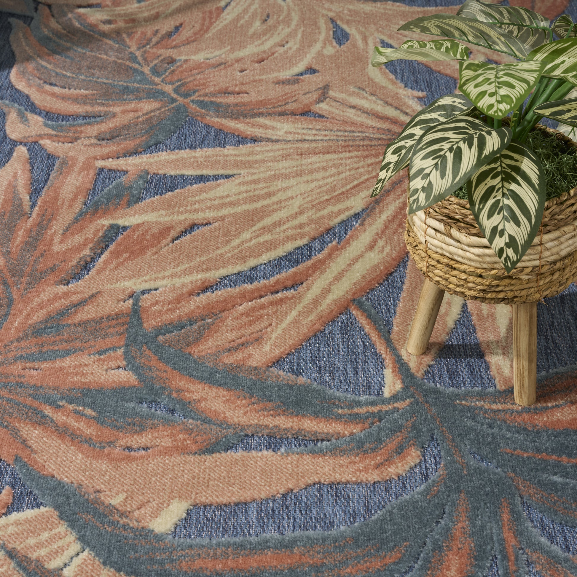 Abstract leaf-pattern rug in peach and blue tones with a green-and-cream variegated plant in a woven basket on a wooden stand.