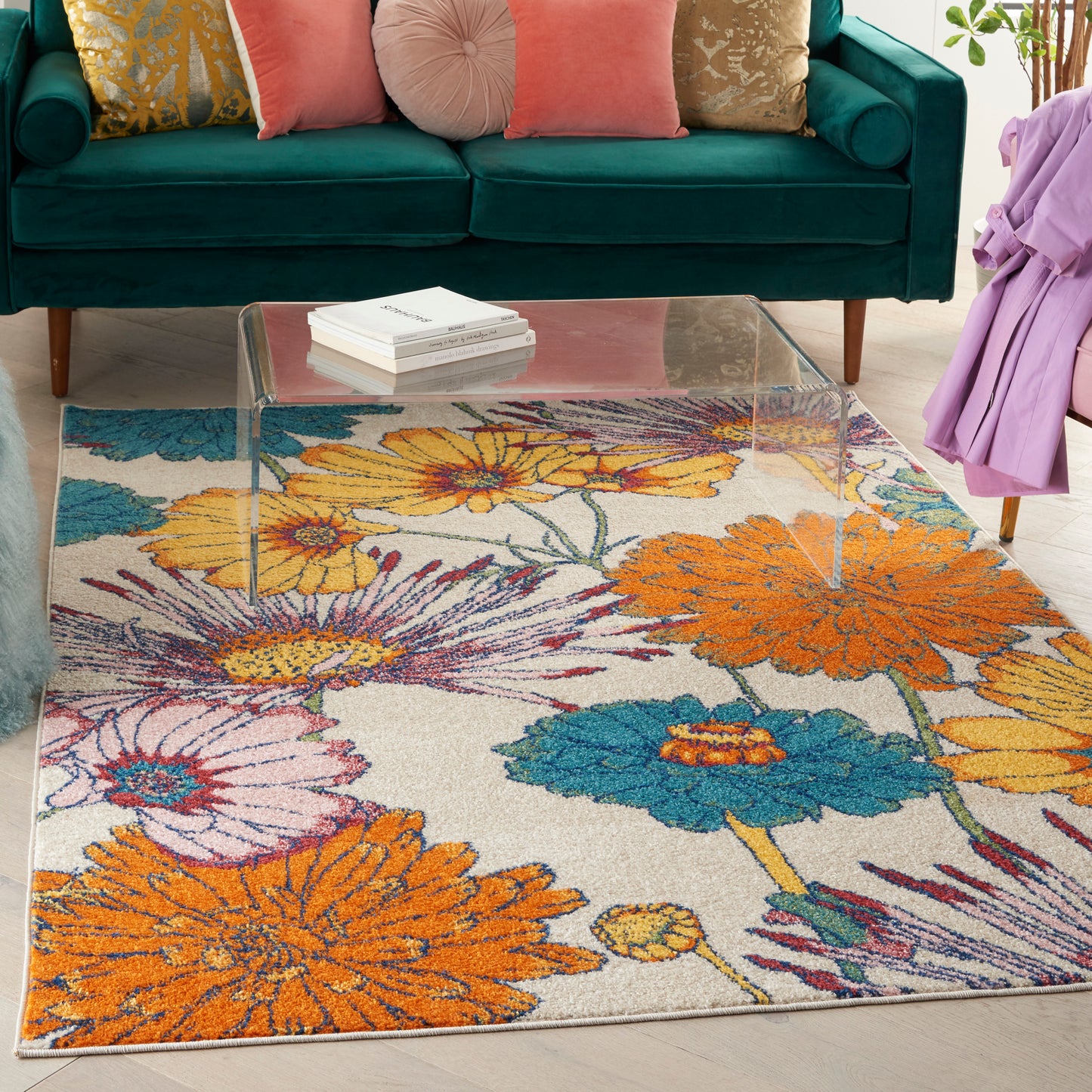 Teal velvet sofa with pink cushions, clear glass coffee table, vibrant floral rug in orange, yellow, blue.
