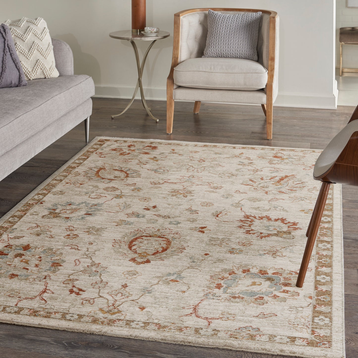 Beige traditional area rug with rust and blue floral motifs and border, in a living room with grey sofa and wooden armchair.