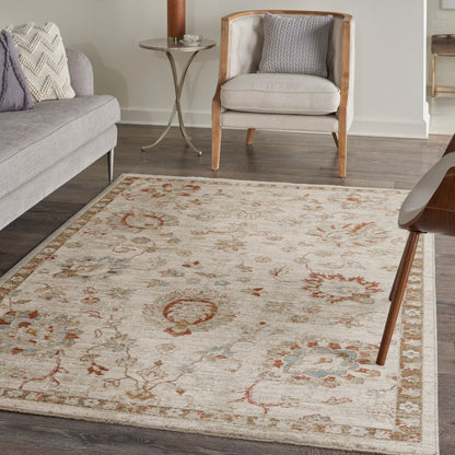 Beige traditional area rug with rust and blue floral motifs and border, in a living room with grey sofa and wooden armchair.
