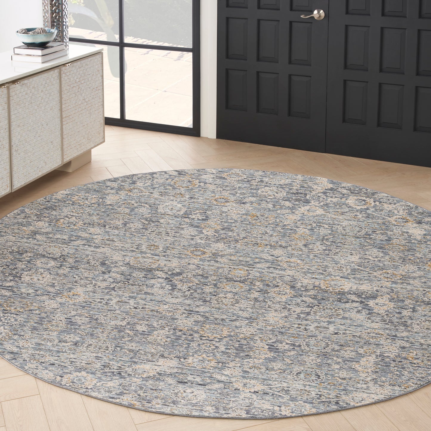 Round blue-gray area rug with ivory-gold pattern in entryway, near black paneled doors and light wood floor.