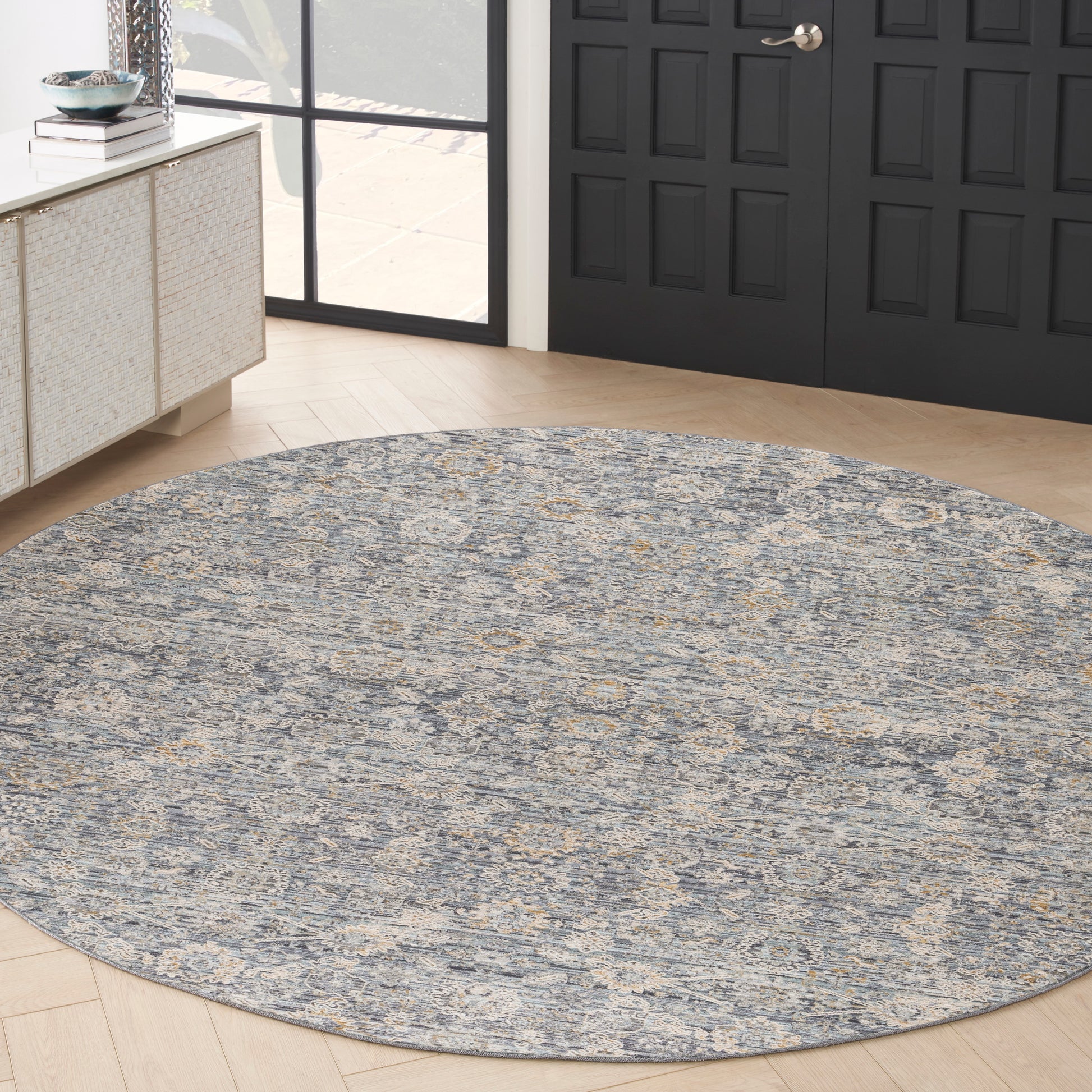 Round blue-gray area rug with ivory-gold pattern in entryway, near black paneled doors and light wood floor.