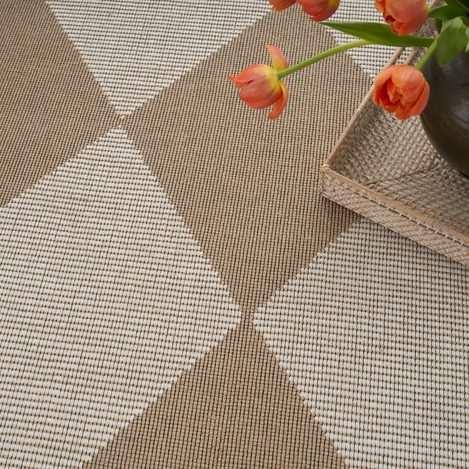 Woven beige and tan diamond-pattern rug with a wicker tray and orange tulips in a vase.