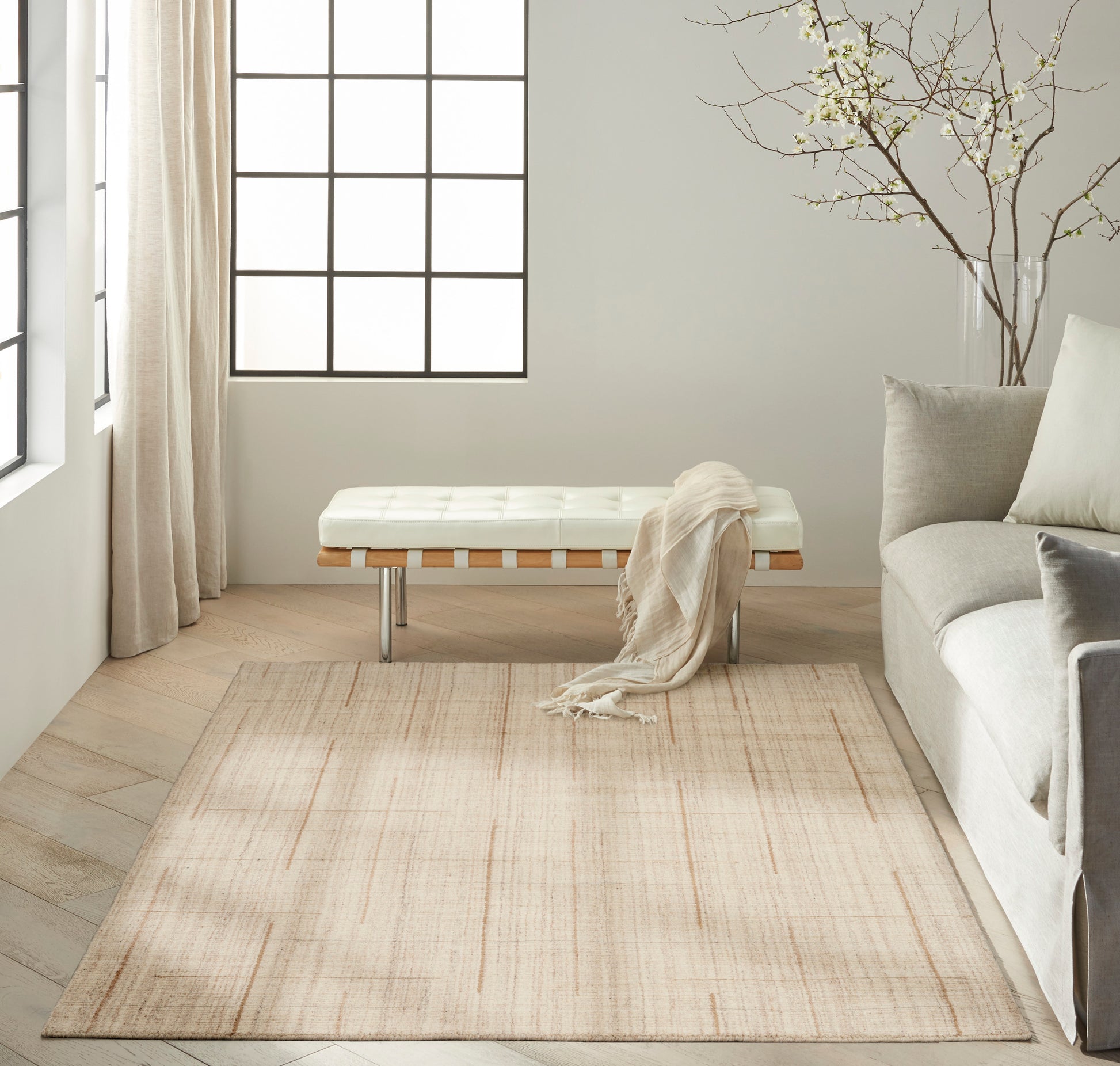 Bright minimalist living room with tufted white bench, pale beige rug, light sofa, sheer curtains, and a bare branch decor.