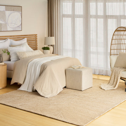 Beige rug in a neutral bedroom with a wooden slat headboard, linen bedding, a woven ottoman, sheer-curtain windows, and a rattan chair.