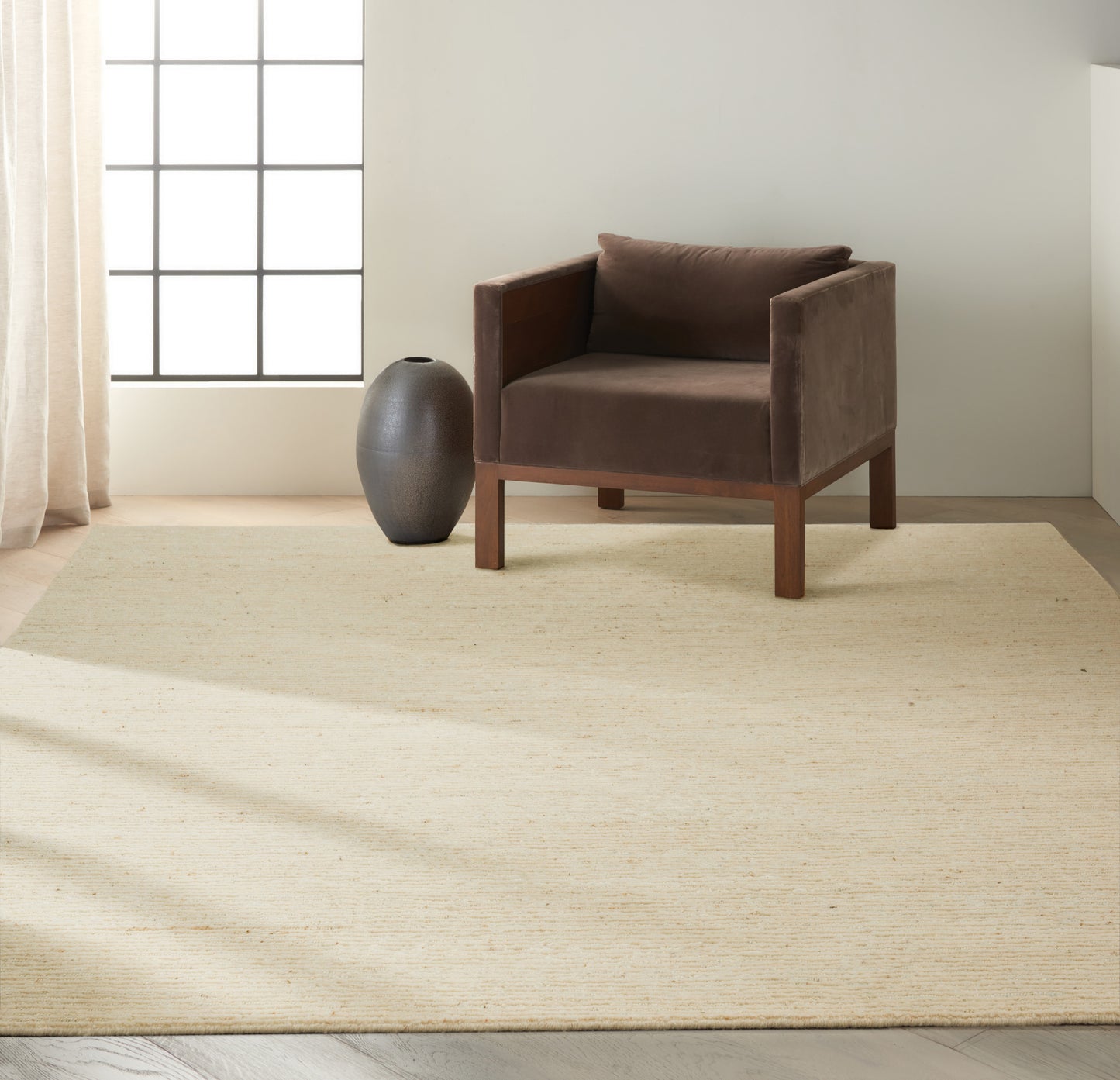 Brown velvet armchair with wooden frame on a beige woven rug, next to a dark ceramic vase in a sunlit minimalist room.