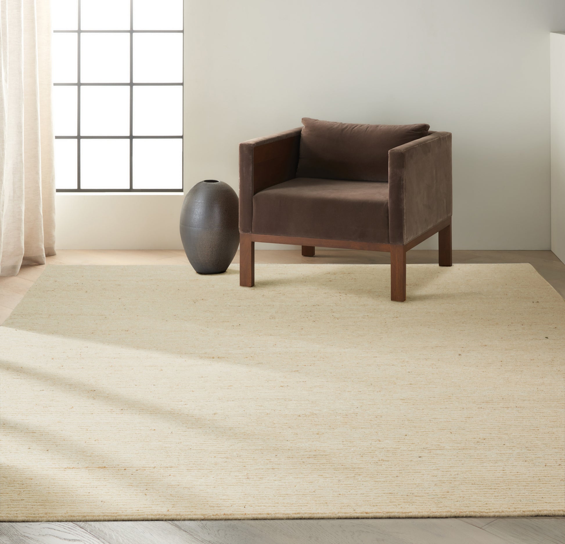 Brown velvet armchair with wooden frame on a beige woven rug, next to a dark ceramic vase in a sunlit minimalist room.