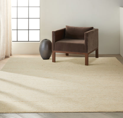 Brown velvet armchair with wooden frame on a beige woven rug, next to a dark ceramic vase in a sunlit minimalist room.