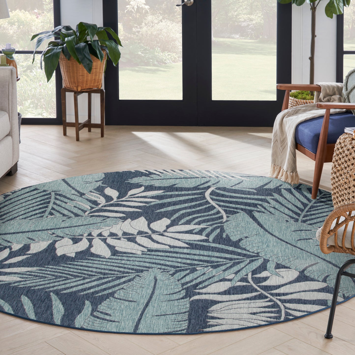 Round teal and navy botanical outdoor rug in a bright patio seating area with wicker furniture, a potted plant, and glass doors.