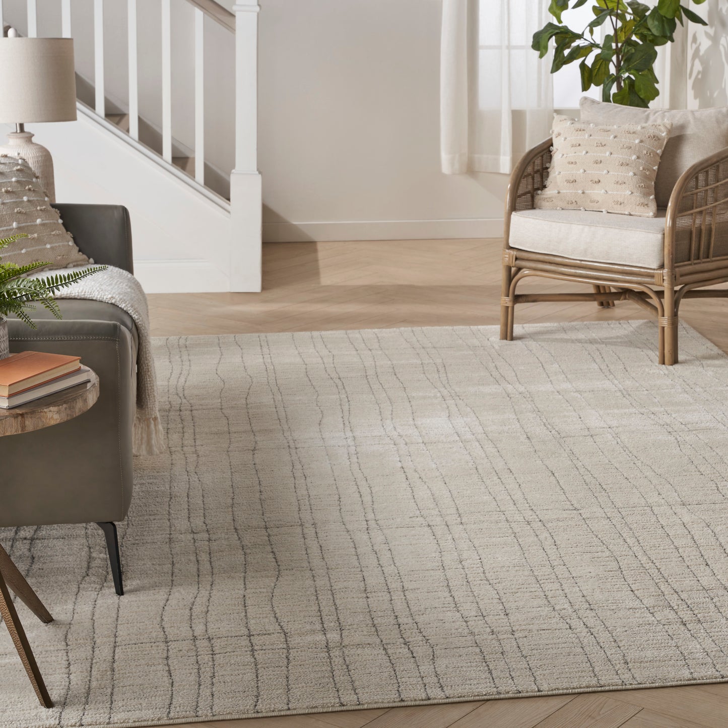 Bright living room with a cream-gray patterned area rug, gray sofa, wicker chair, plant, and staircase in the background.