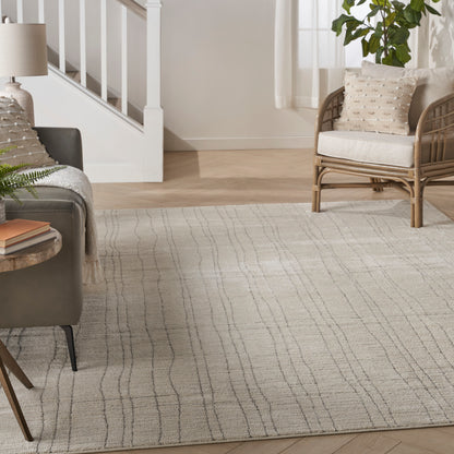Bright living room with a cream-gray patterned area rug, gray sofa, wicker chair, plant, and staircase in the background.