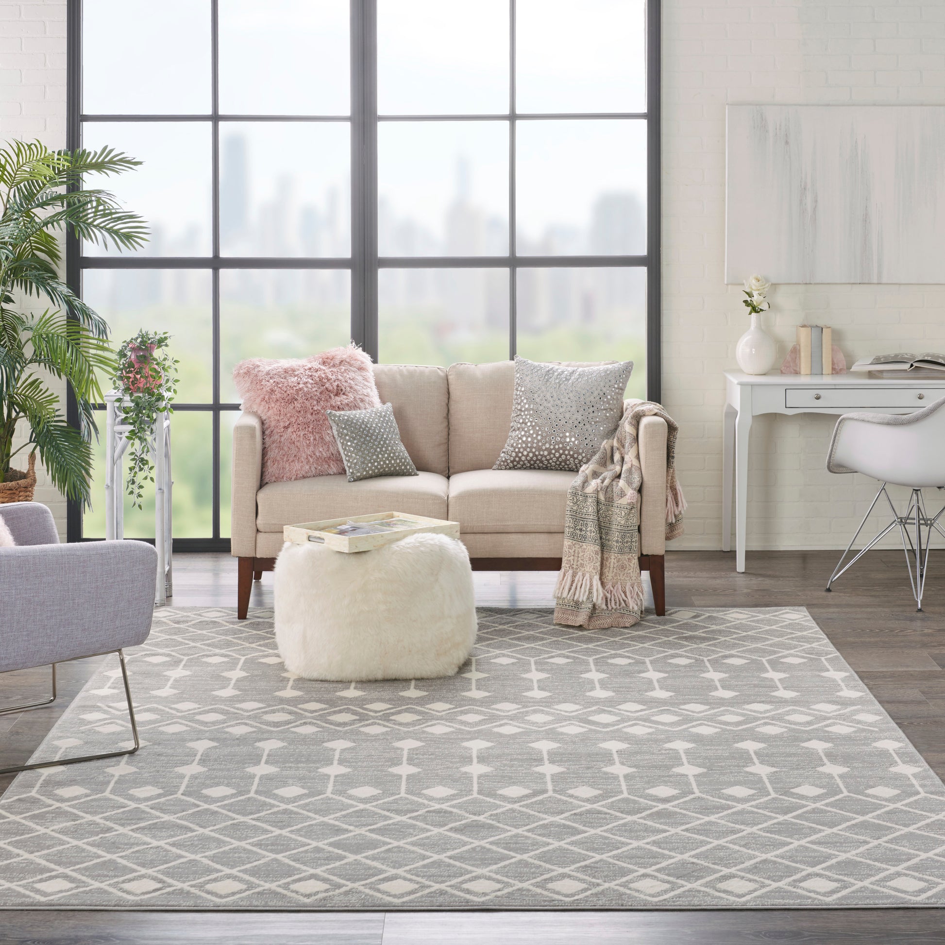 Bright living room with grey geometric rug, beige sofa with pink/gray cushions, fluffy white pouf, plant, and a white desk.