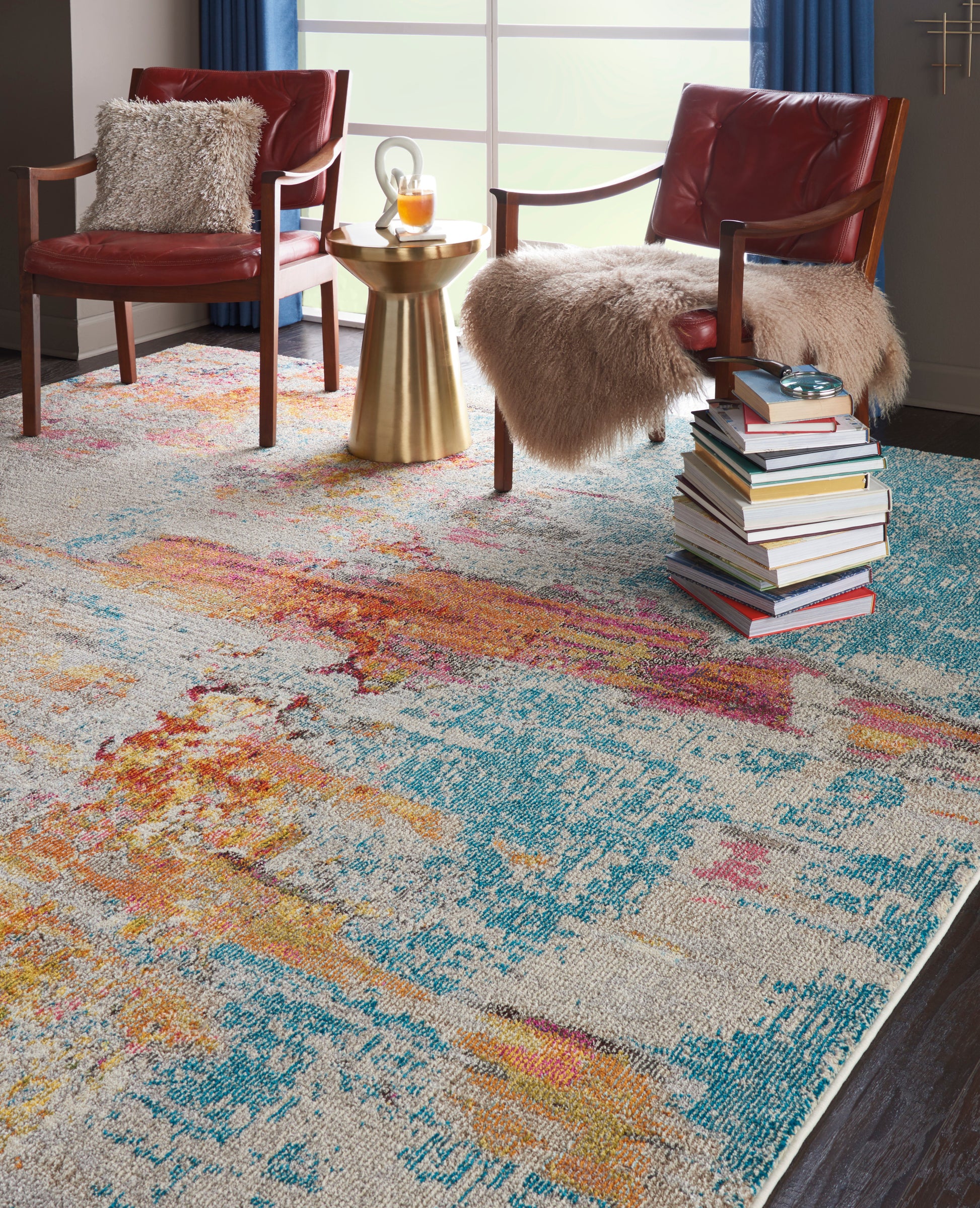 Vibrant abstract area rug in teal, orange, yellow & pink in a living room, with red leather chairs, fur throws and a gold side table; size 7' x 10'.