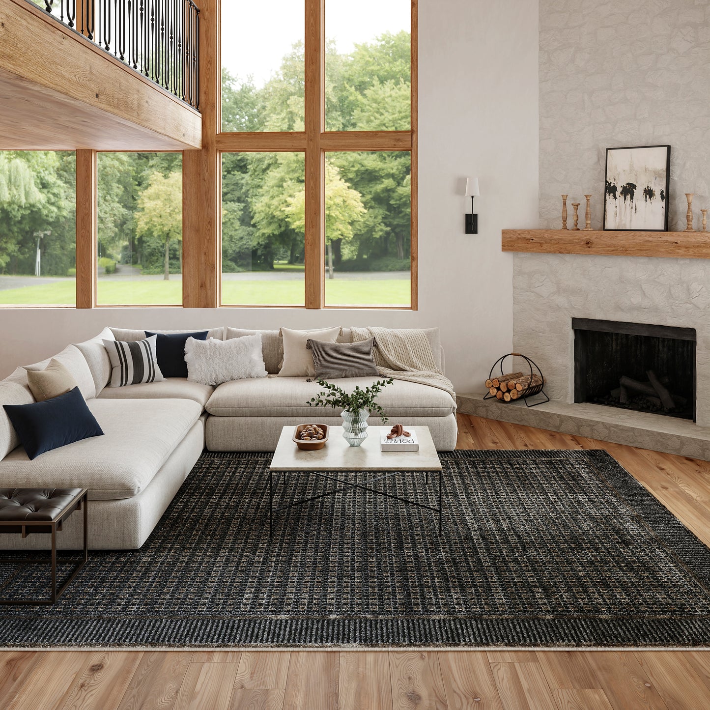 Bright living room with large wood-framed windows, beige sectional, white coffee table, dark rug, and stone fireplace.