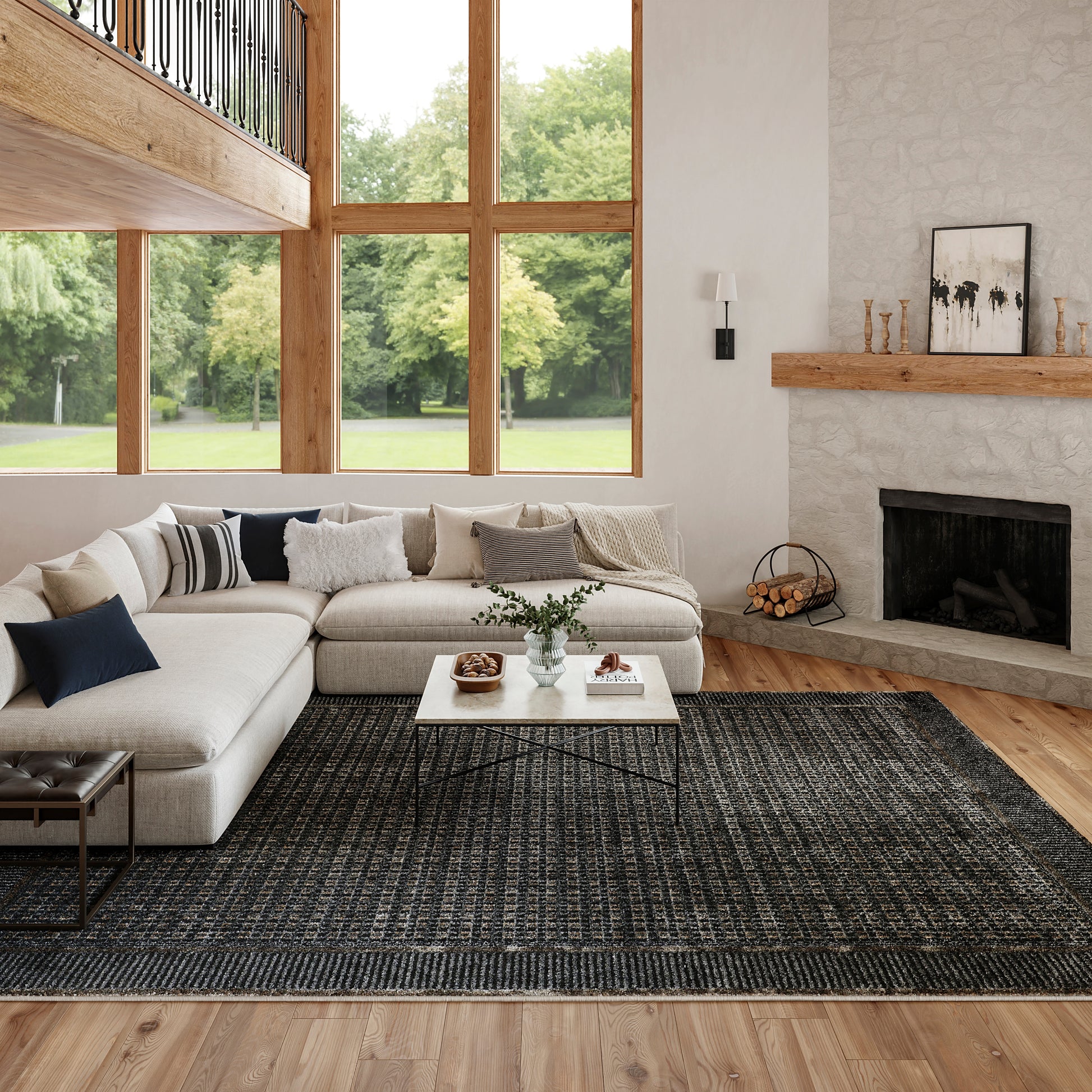 Bright living room with large wood-framed windows, beige sectional, white coffee table, dark rug, and stone fireplace.