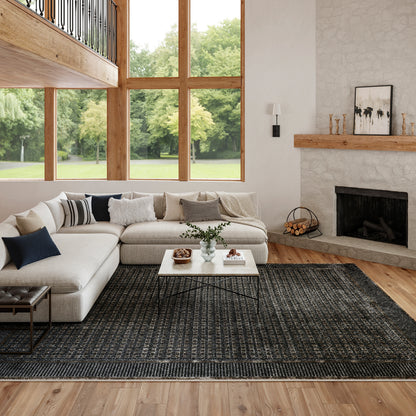 Bright living room with large wood-framed windows, beige sectional, white coffee table, dark rug, and stone fireplace.