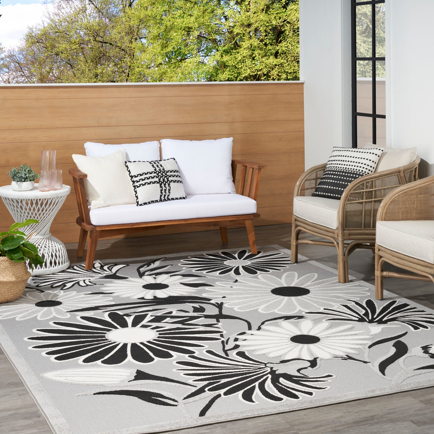 Outdoor seating area with light wood sofa, beige wicker chairs, white cushions, black-and-white floral rug.
