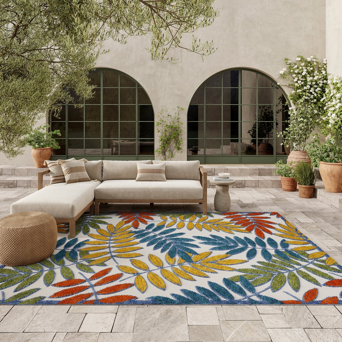 Outdoor patio with a colorful leaf-print rug in yellow, blue, orange, and green, beige sectional sofa, and potted plants.