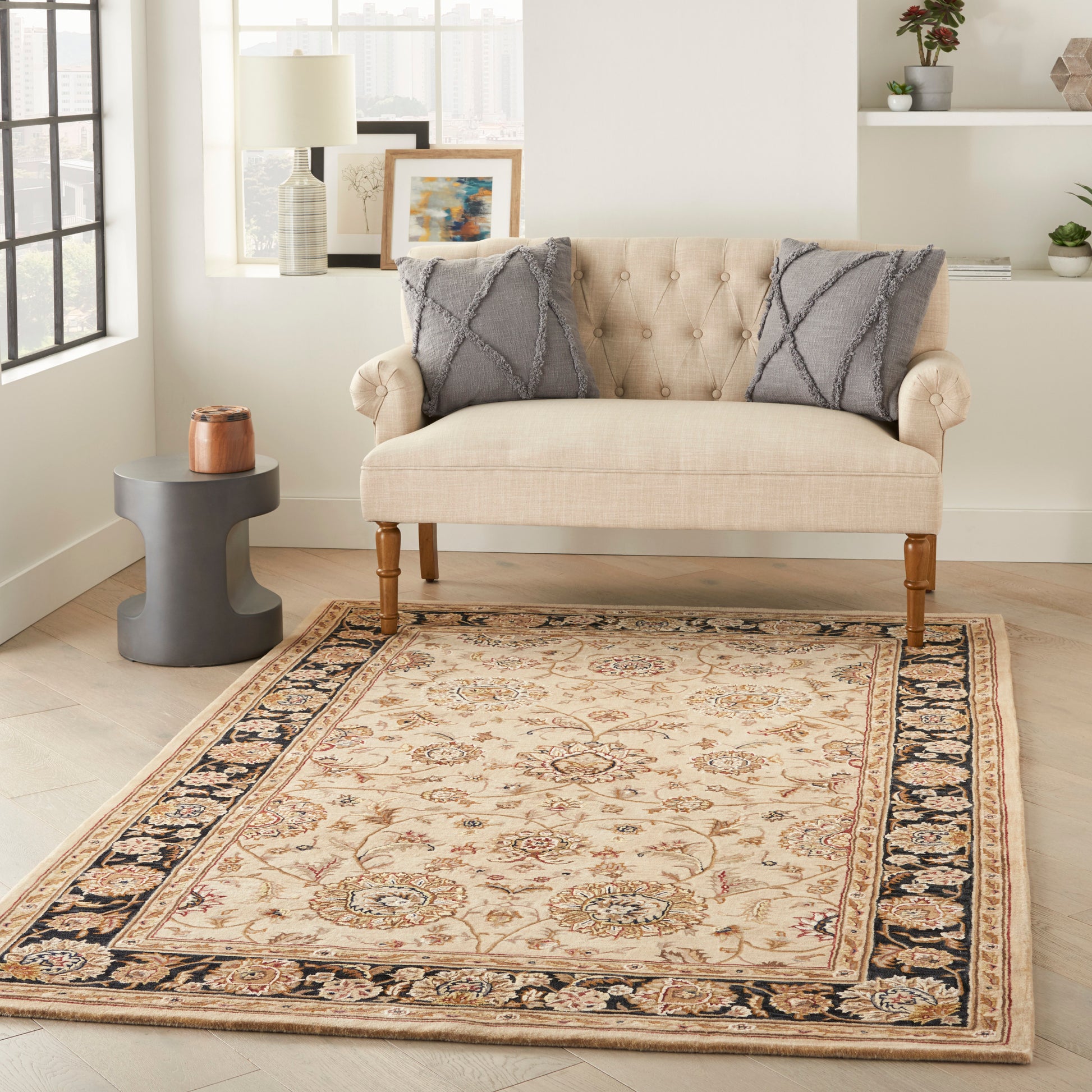 Beige tufted loveseat with wooden legs, two gray diamond pillows, ornate beige area rug with border in a cozy living room.