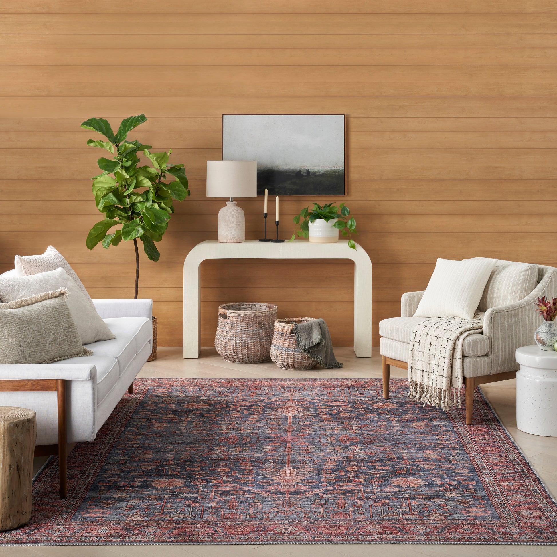 Living room with wood-paneled wall, white curved console, lamp, framed art, plants, baskets, and a red-blue patterned rug.
