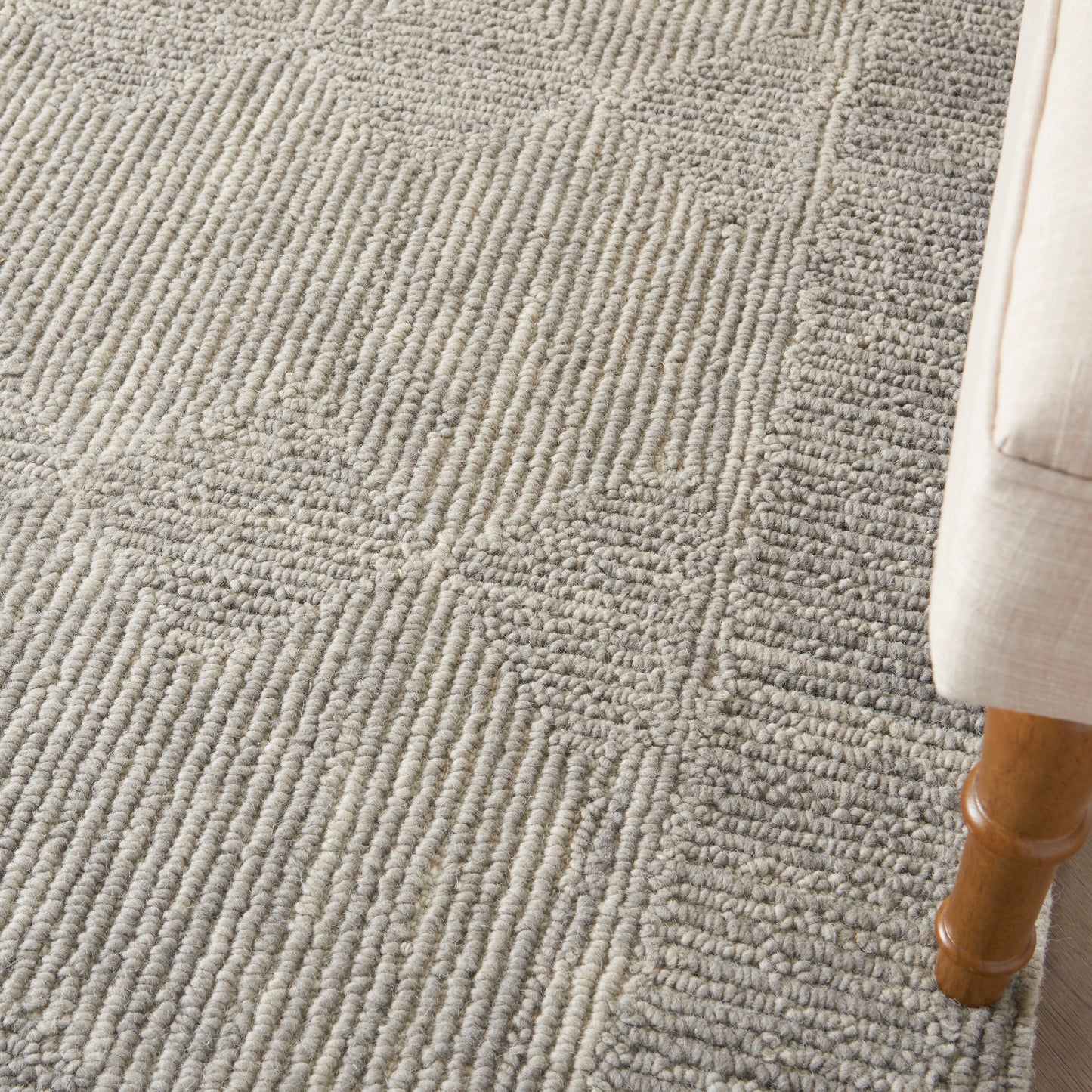 Gray textured rug with looped pile and geometric block pattern beside a beige sofa leg.