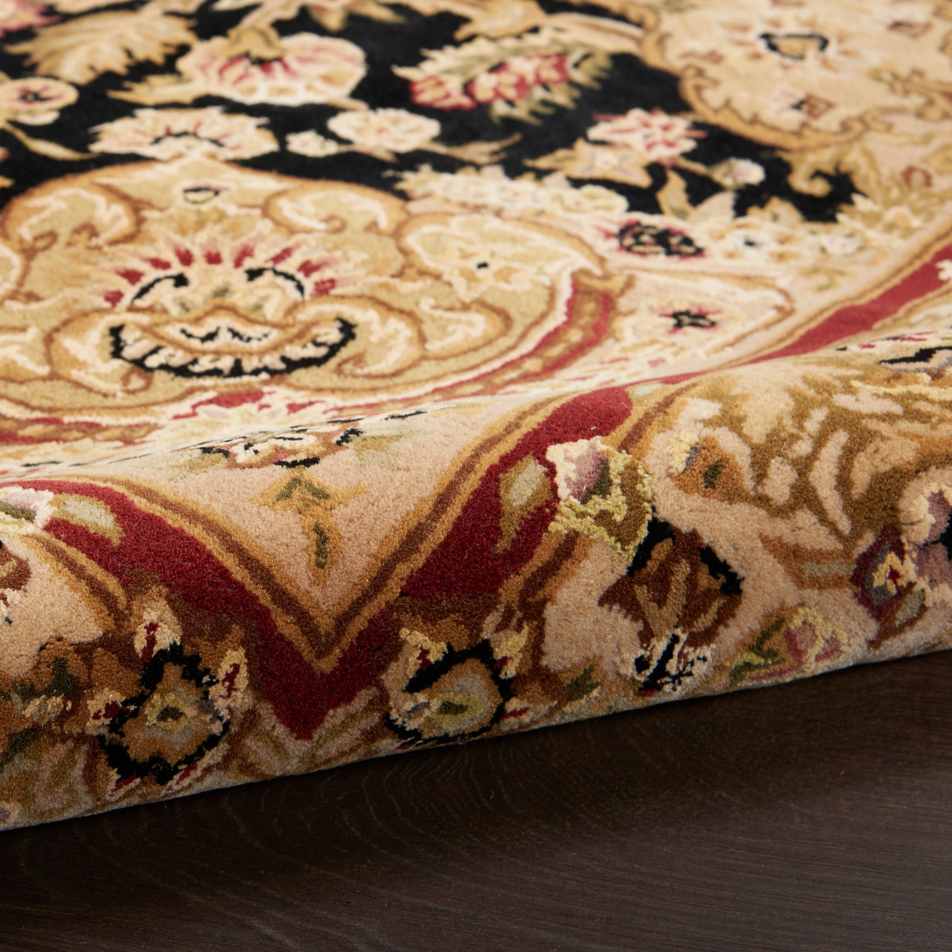 Close-up of ornate Persian-style rug with gold, beige, black and red floral damask patterns; thick pile on a dark wood floor.