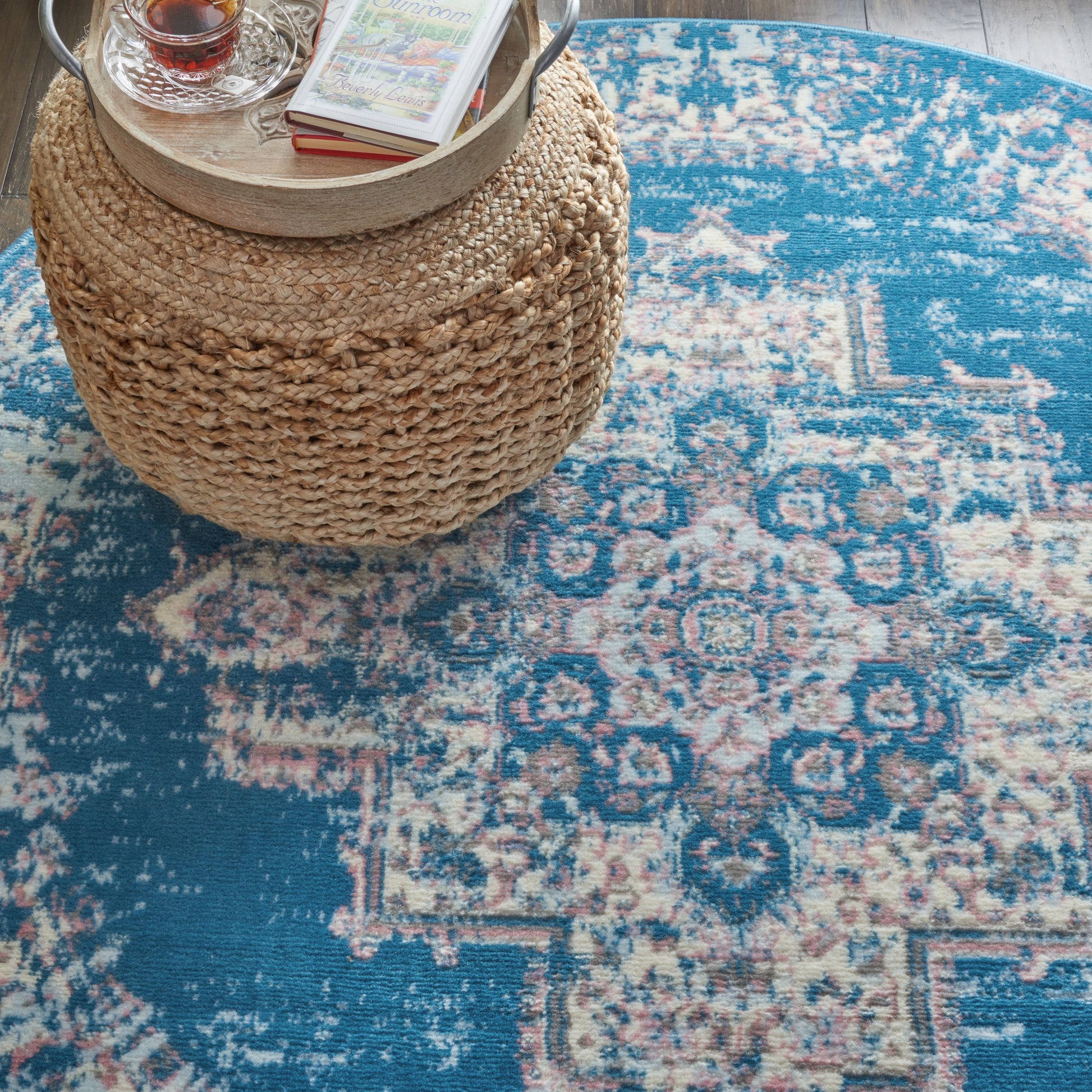 Round woven rattan ottoman with a wooden tray, glass teacup and books, on a blue oriental rug.