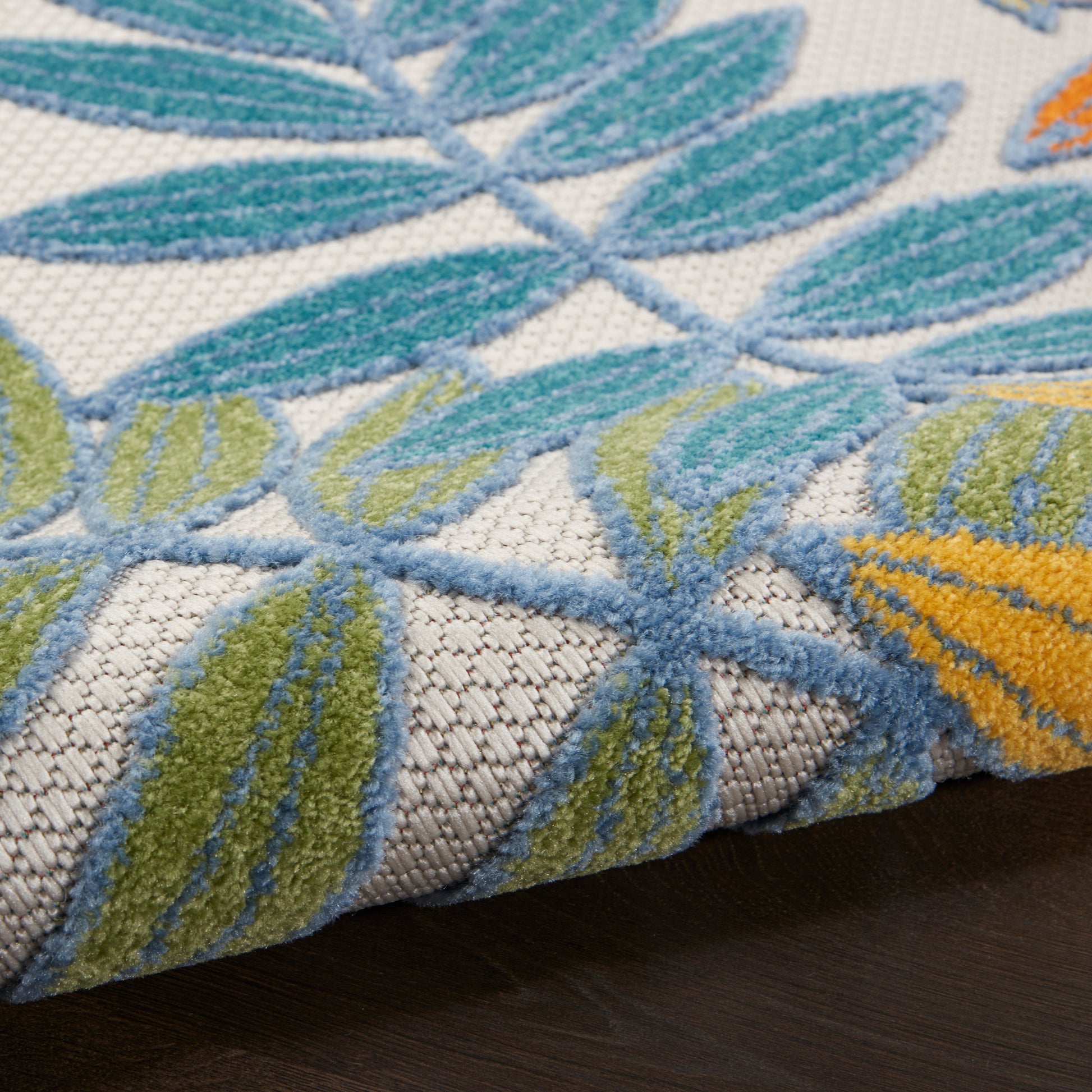Close-up of a tufted rug with blue, green & yellow leaf motifs on a light beige background; folded edge.