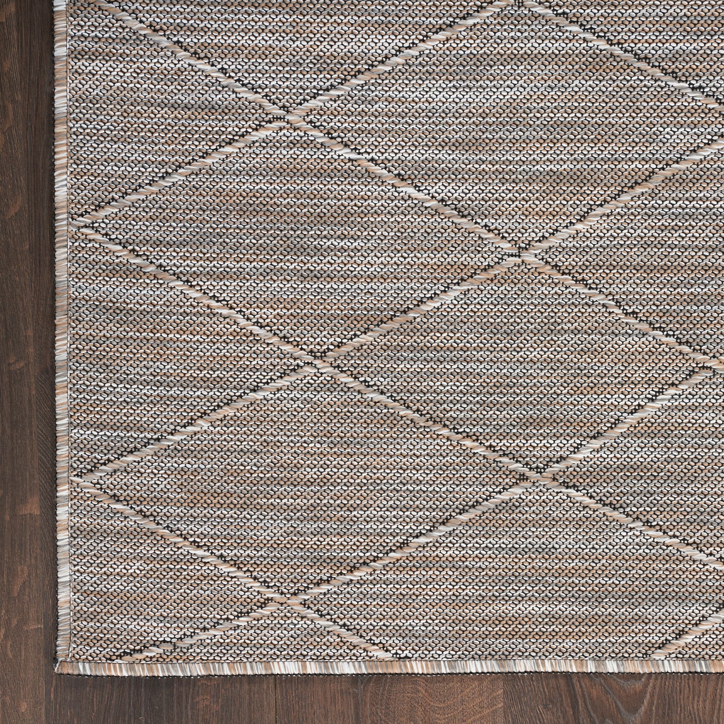 Beige-taupe woven rug with diamond lattice pattern, textured surface, fringe edging on dark wood floor.