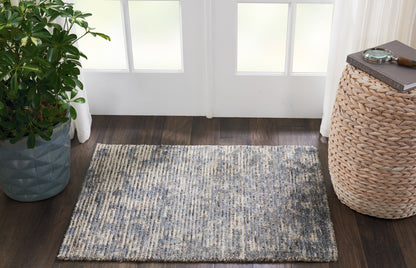 Rectangular blue-gray woven area rug in an entryway with a potted plant on the left and a wicker side table on the right.