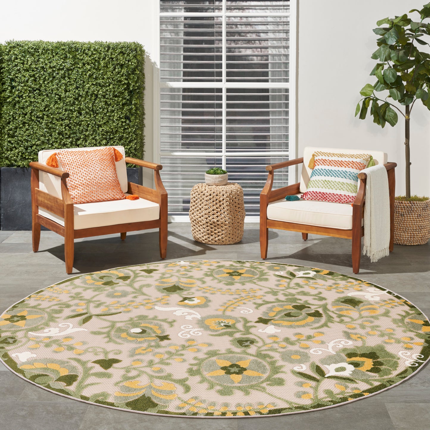 Outdoor patio: round beige-green floral rug, mid-century wooden armchairs with colorful cushions, woven side table.