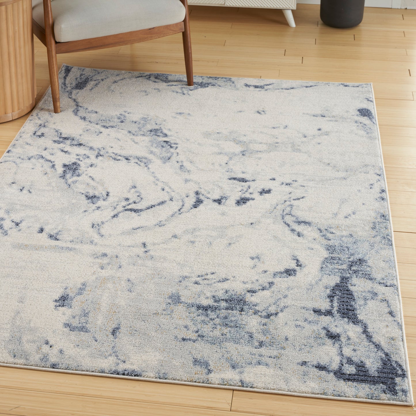 Light cream rectangular area rug with blue marbled abstract pattern on wooden floor beside a chair.
