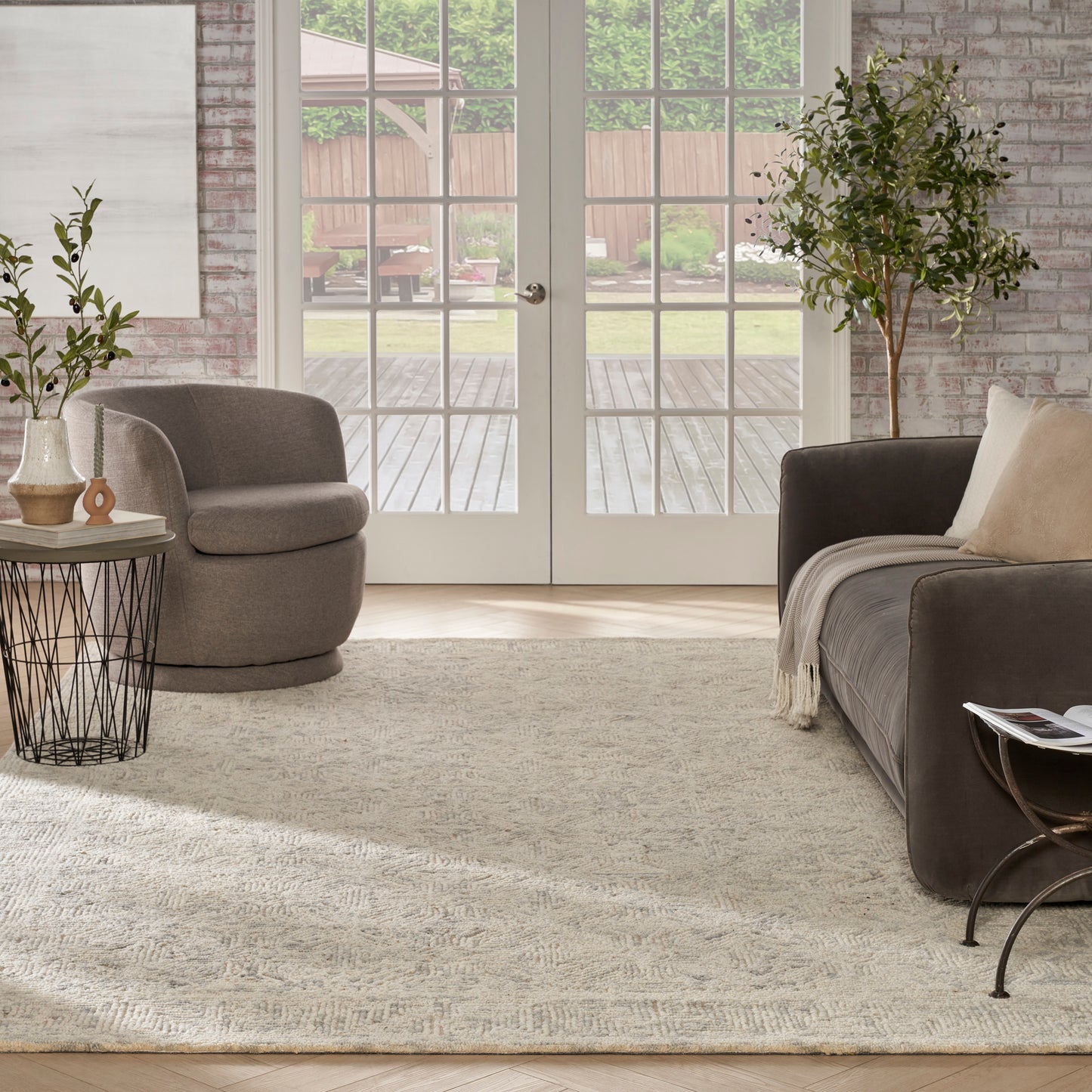 Cozy living room with beige rug, gray round armchair, dark sofa, throw pillows, metal wire side table, potted tree, doors.