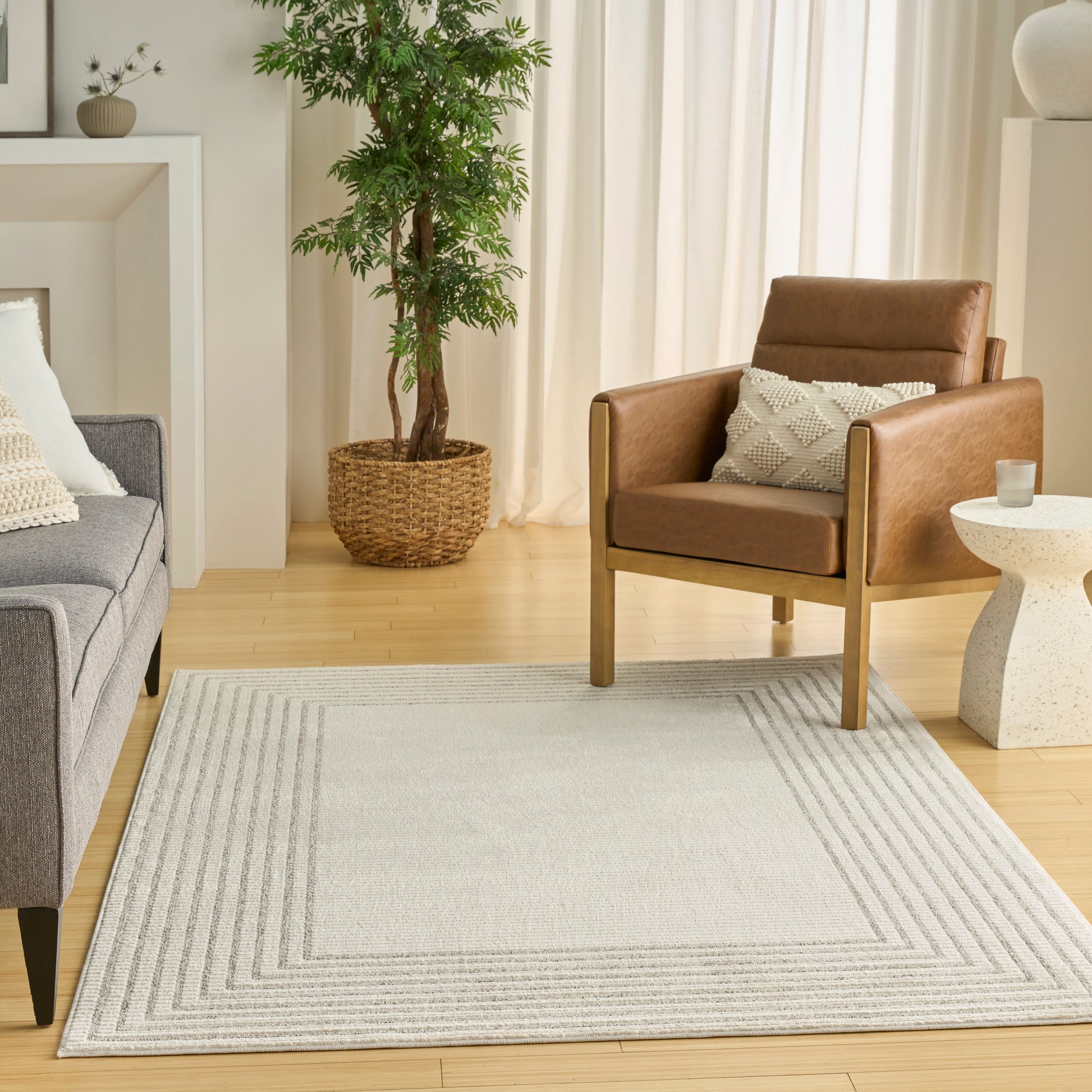 Bright living room scene with grey sofa, brown leather armchair, light geometric rug, potted plant, and white curtains.