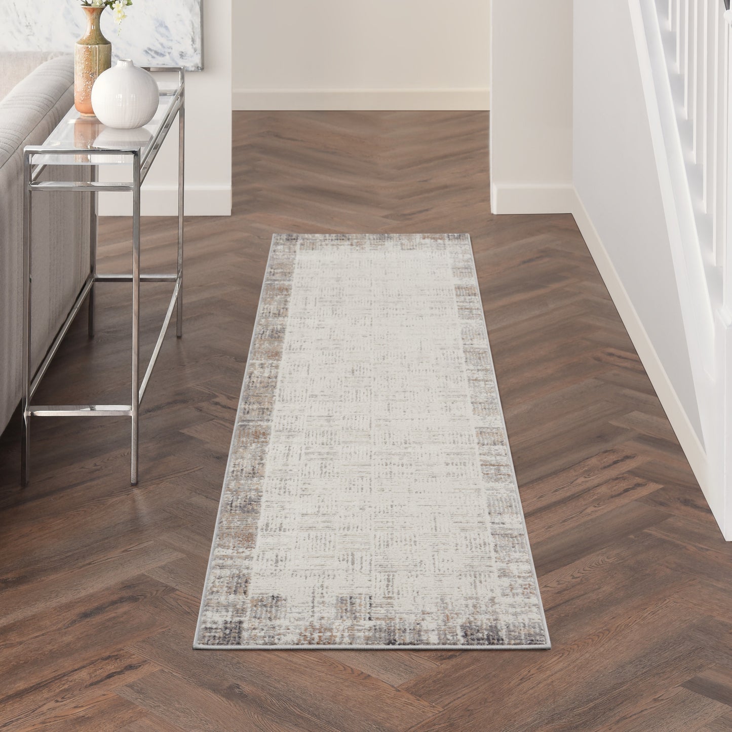 Light beige distressed runner rug with a grey border in a dark herringbone hallway; chrome-framed glass console table.