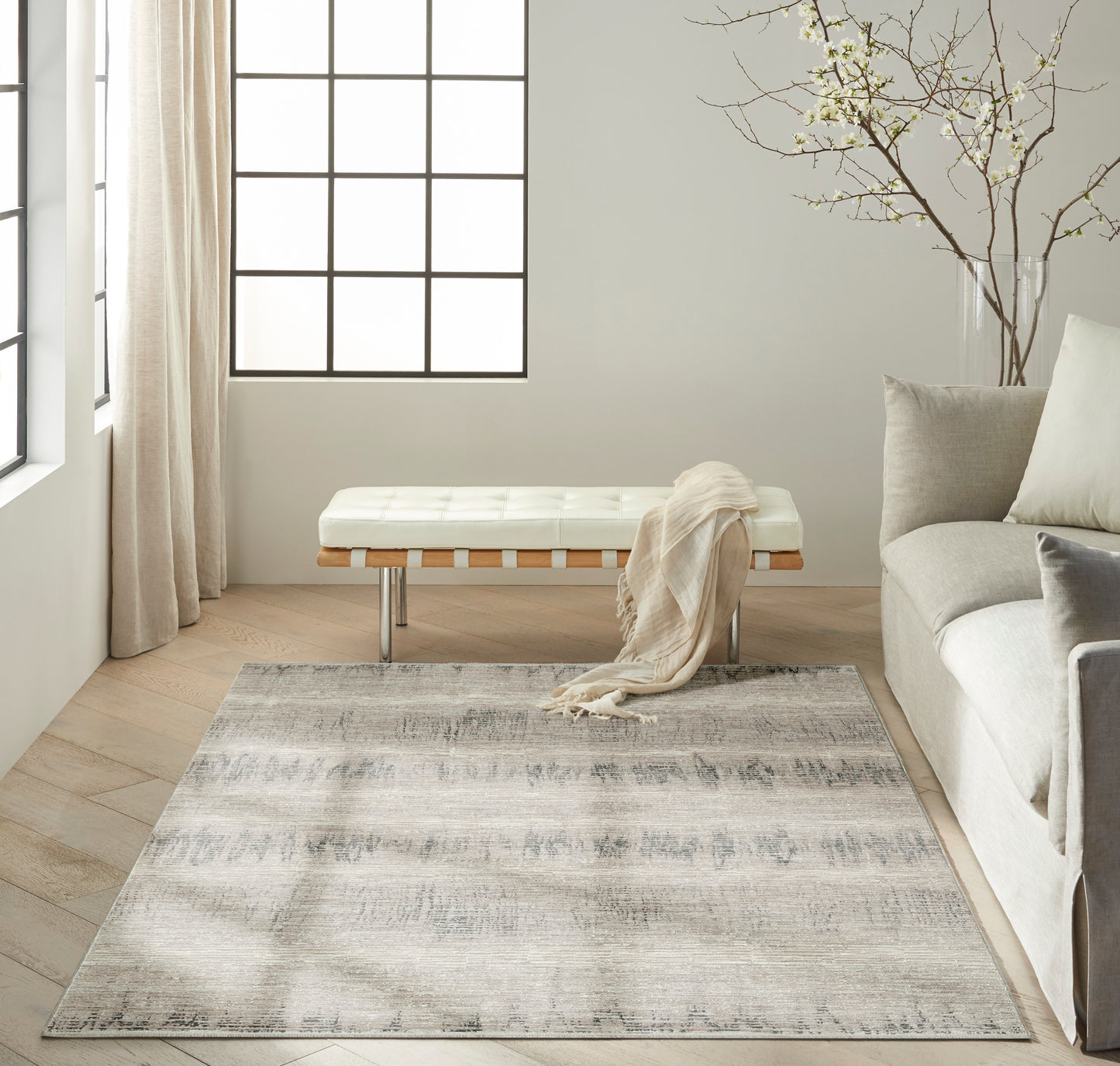 Neutral living room with cream tufted bench, beige throw, gray patterned rug, pale sofa, light wood floors, grid window.