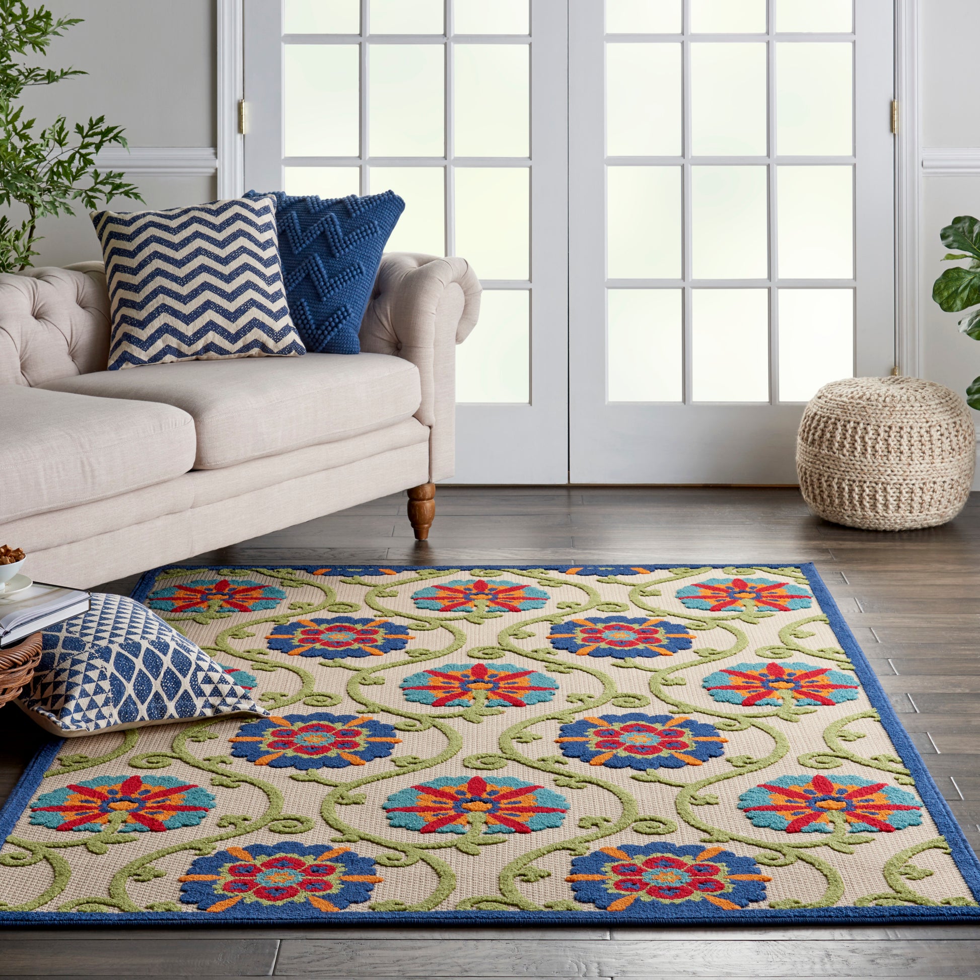 Colorful floral area rug with blue border on beige background & green vines; beige sofa with blue pillows.