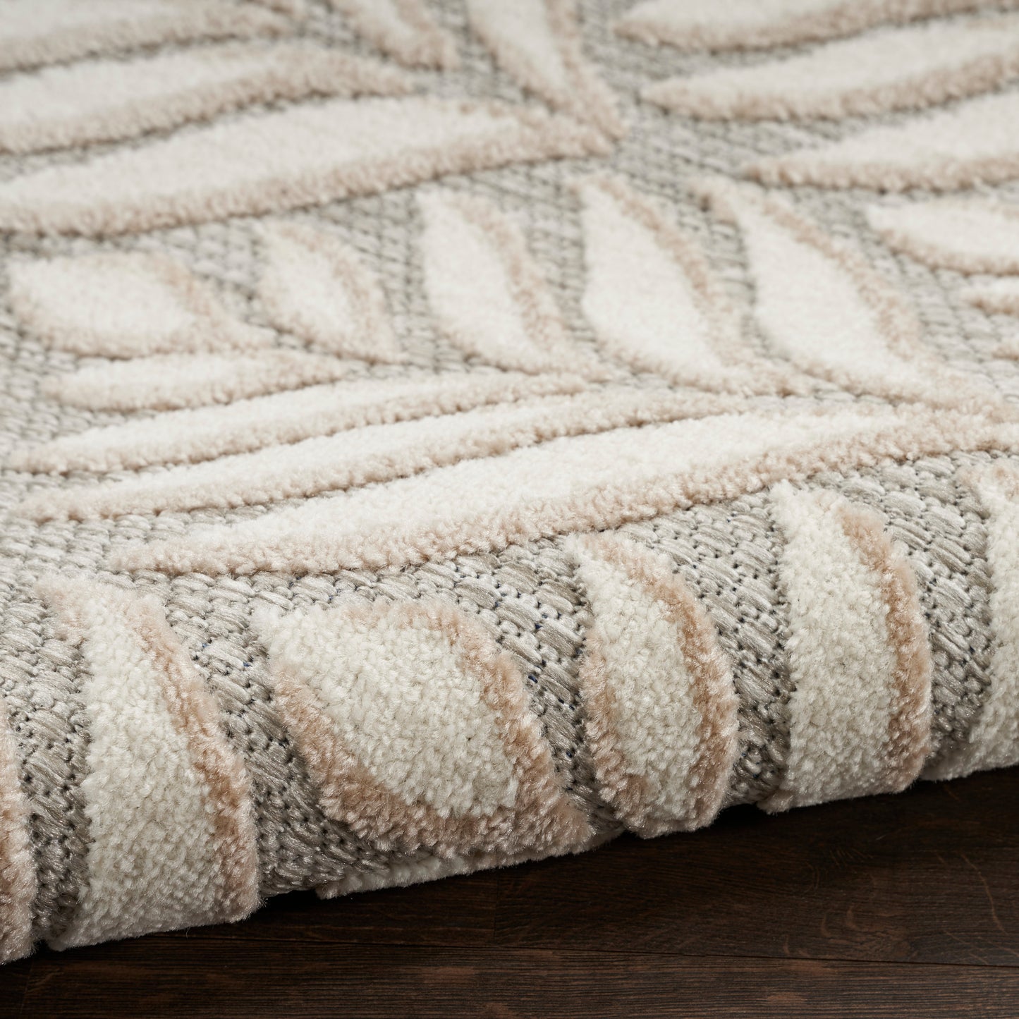 Close-up of a plush beige and gray rug with a raised geometric pattern and rolled edge on dark wood floor.