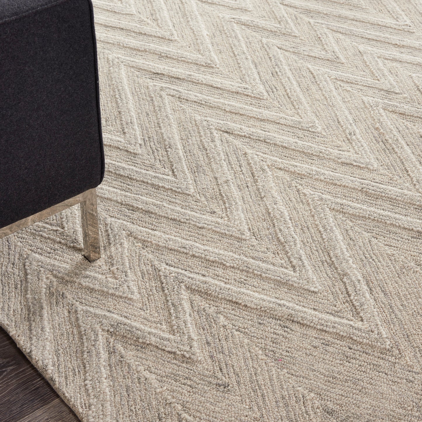 Beige herringbone carpet with a textured pattern; black upholstered chair leg visible on the left and dark wood floor edge.
