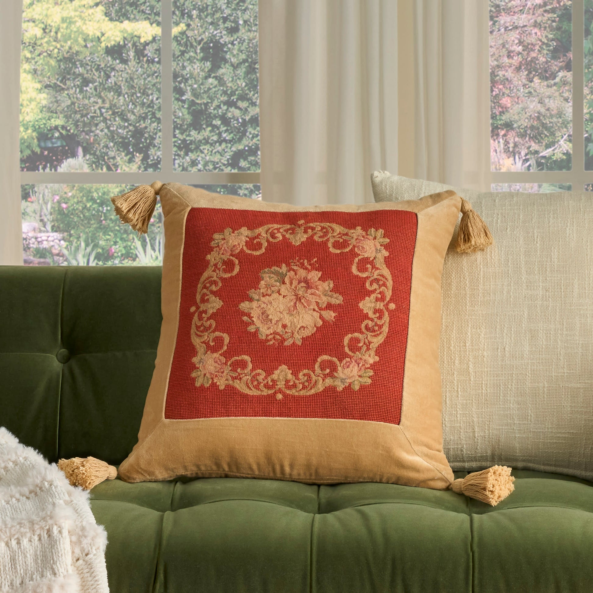 Square throw pillow with tan border, gold tassels, red center and ornate gold embroidery on a green velvet tufted sofa.