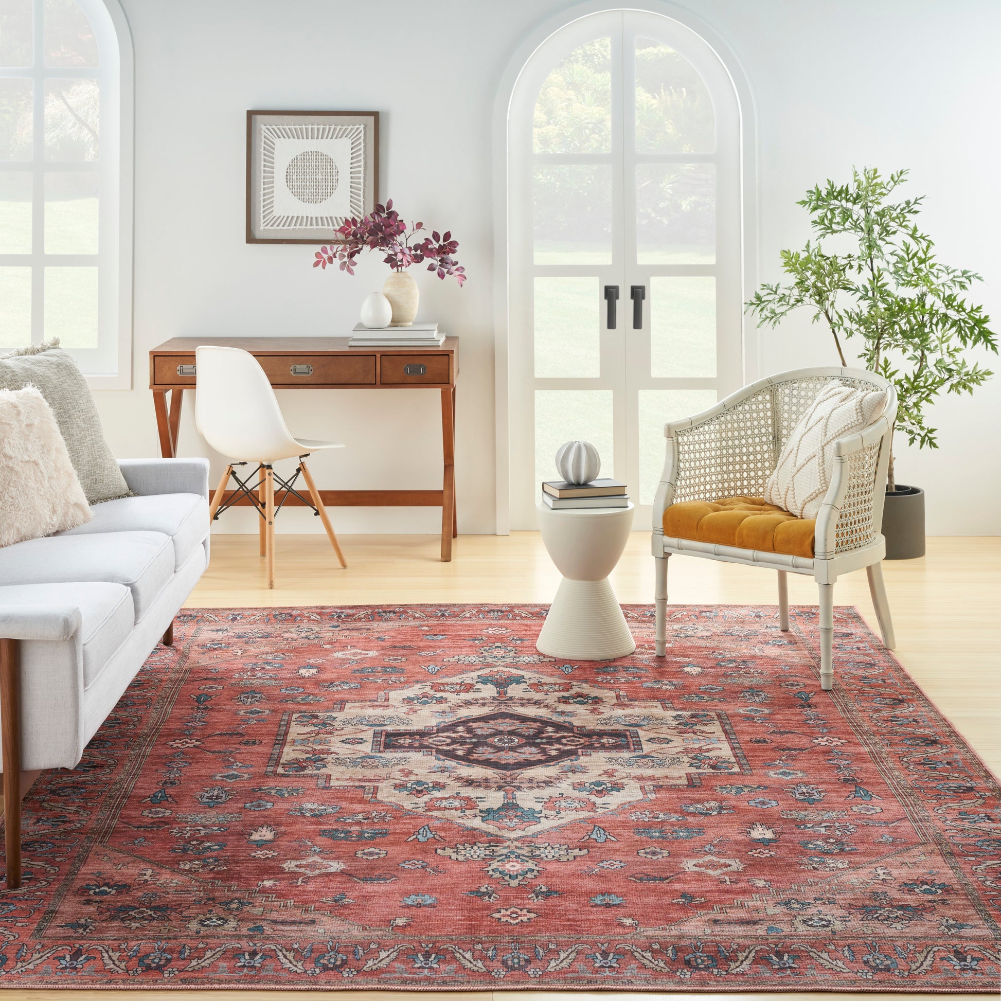 Bright living room with red oriental rug, white sofa, wood desk, wicker chair with orange cushion, arched windows.
