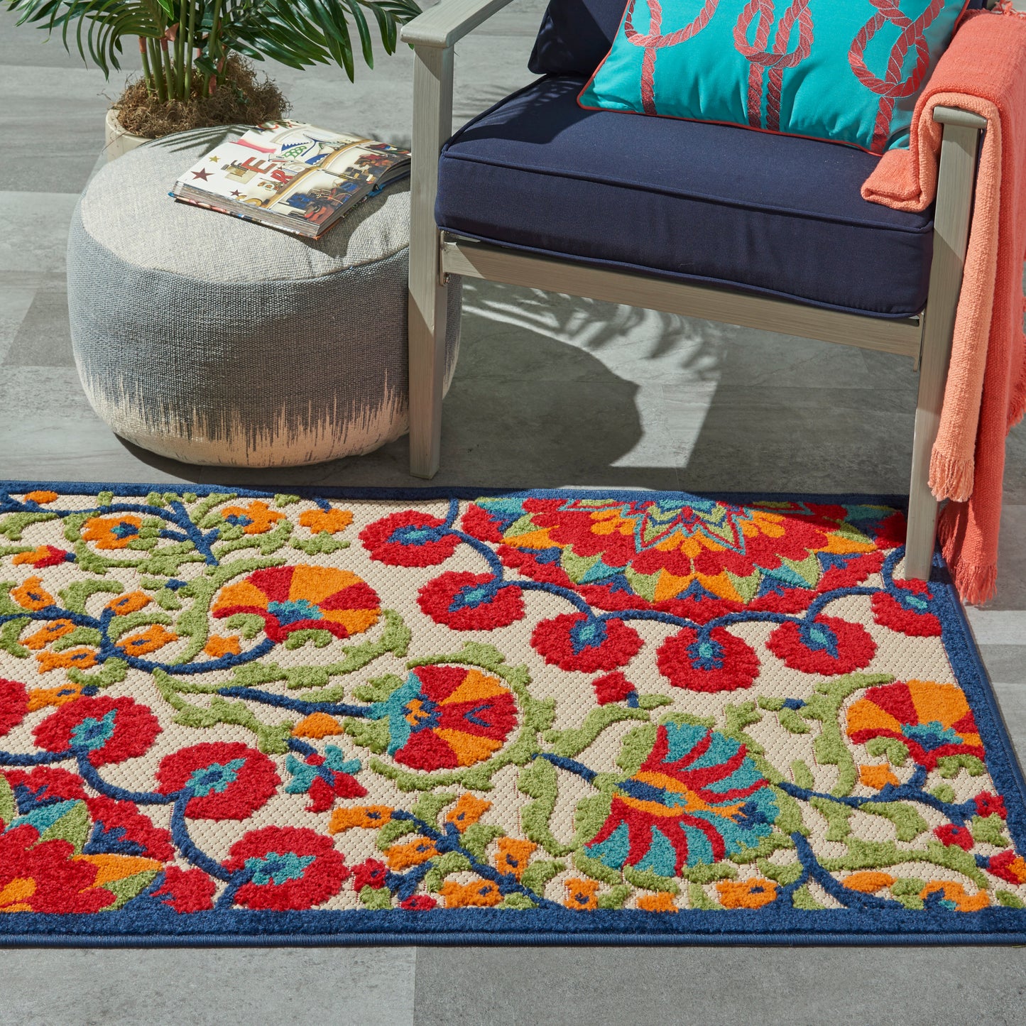 Vibrant outdoor rug with red, blue, green florals; navy chair with turquoise pillow, orange throw; gray pouf; magazines.