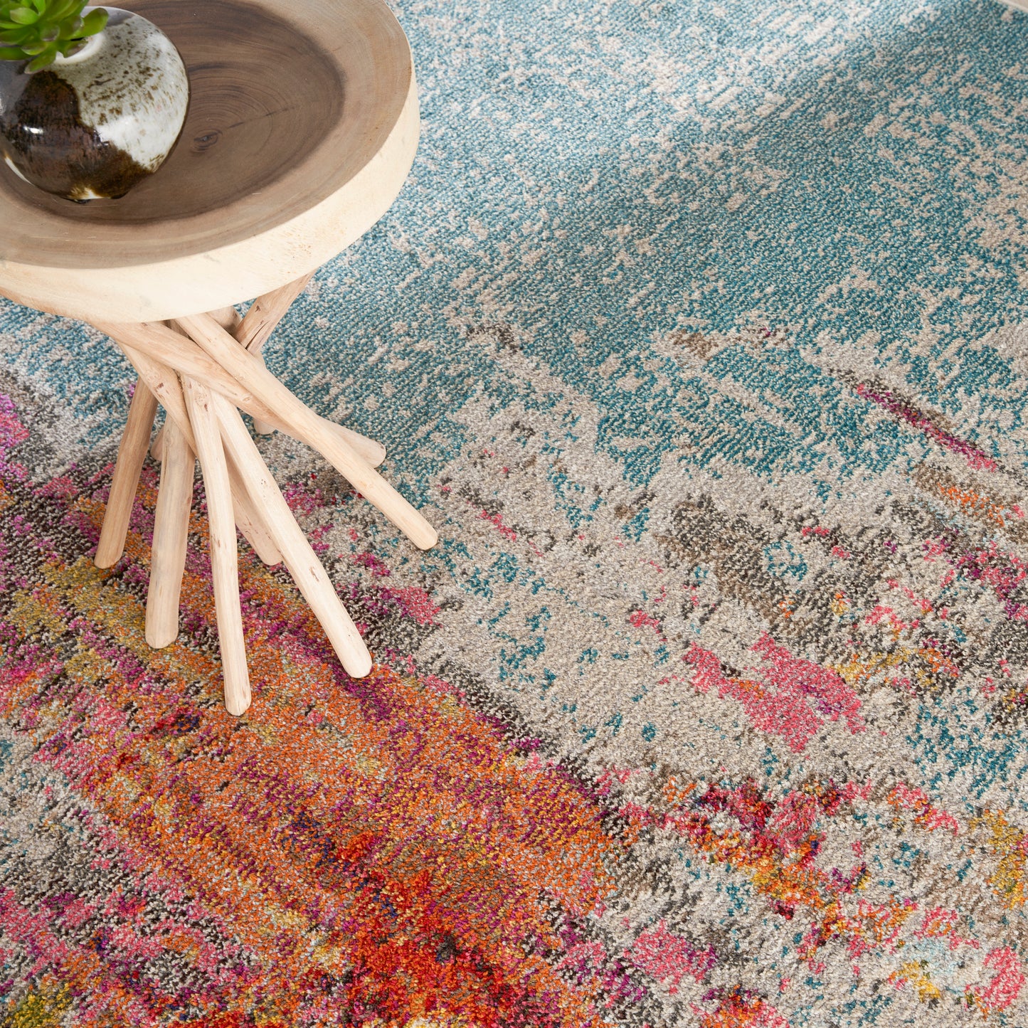 Multicolored abstract area rug in teal, pink, orange tones with a light wood stool and small potted plant.