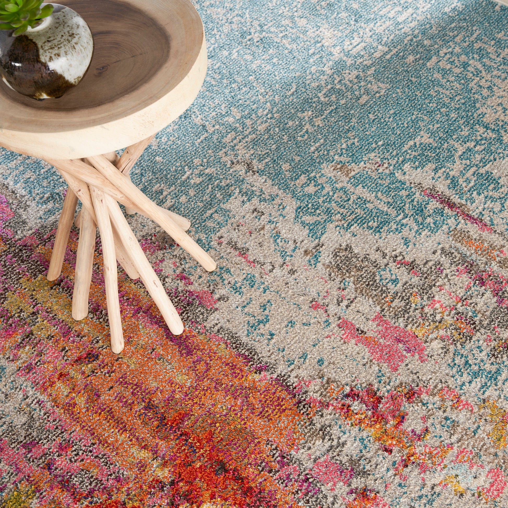 Multicolored abstract area rug in teal, pink, orange tones with a light wood stool and small potted plant.