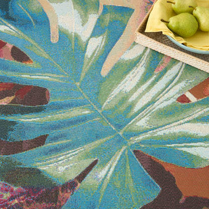 Rectangular tapestry rug with tropical blue-green leaf pattern; top-right wicker tray of pears on a yellow napkin.