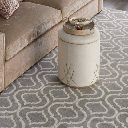 Beige cylinder ottoman with a round metal tray, cup & snacks, on a grey Moroccan-pattern rug beside a tan sofa.
