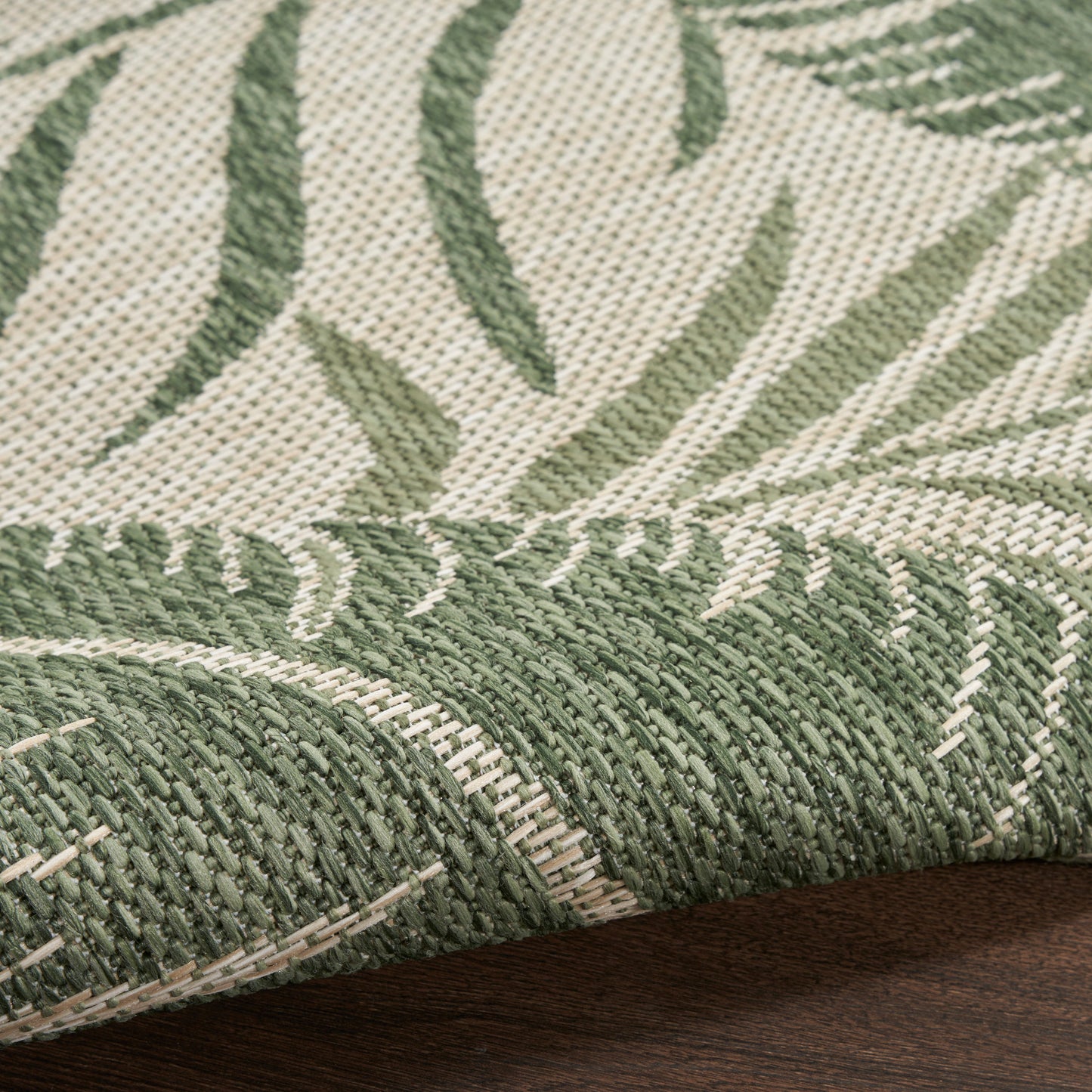 Close-up of green and ivory woven rug with leafy pattern and rolled edge on dark wood surface.